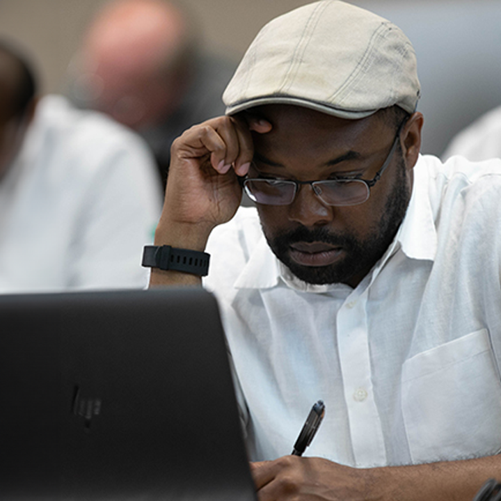A student sitting at a desk, in front of an opened laptop, looking down with a pen in their hand.